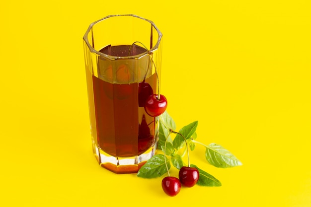 Front view of cherry juice inside long glass with green leaves on the yellow surface