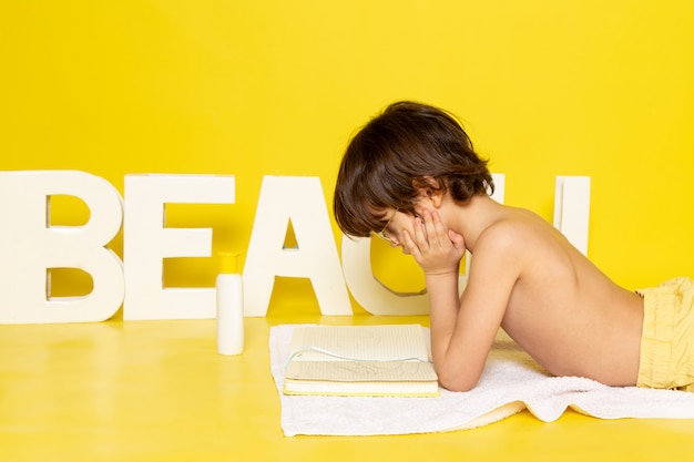 Ragazzo del bambino di vista frontale che si siede sull'asciugamano bianco con la spiaggia di parola sulla scrivania gialla