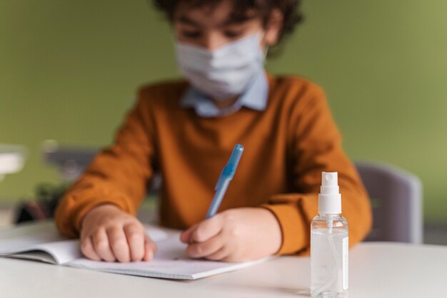 Вид спереди ребенка с медицинской маской в классе с бутылкой дезинфицирующего средства для рук на столе