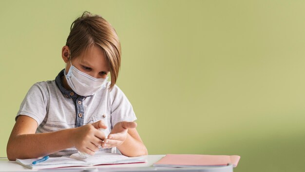 Front view of child with medical mask disinfecting hands in class with copy space