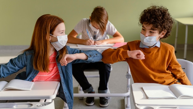 Front view of children with medical masks doing the elbow salute in class