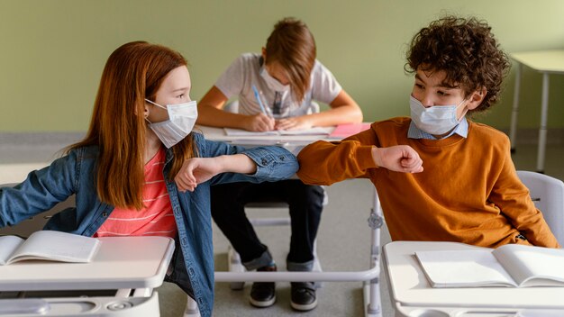 Вид спереди детей в медицинских масках, салютующих локтями в классе