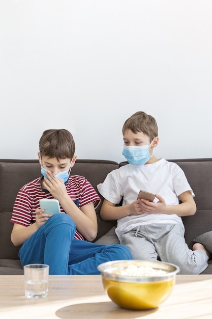 Front view of children with medical masks playing on smartphones