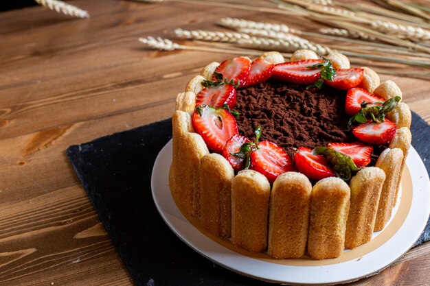 スライスした赤いイチゴビスケットで飾られた正面のチョコレートケーキは、茶色の机の上の白い皿の中のおいしい丸い甘いクッキー菓子