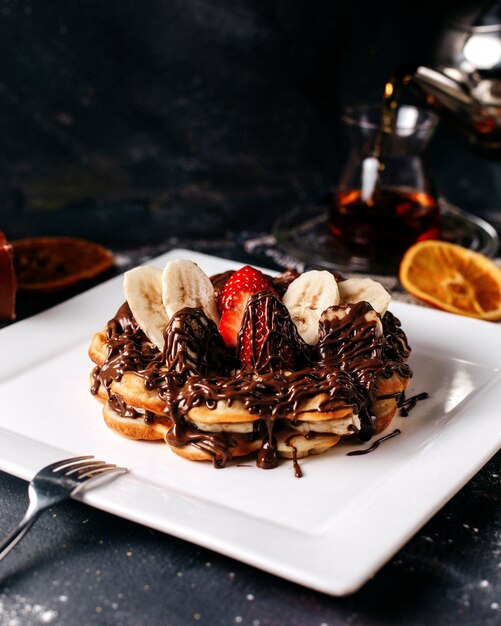 Front view choco pancakes with bananas and red strawberries inside white plate on the bright desk