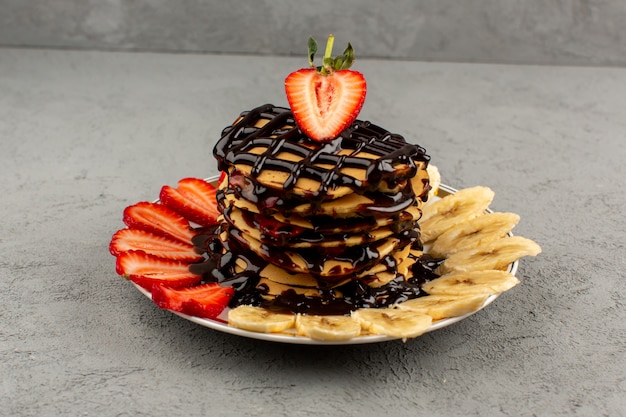 front view choco pancakes with red sliced strawberries and bananas inside plate on the grey floor