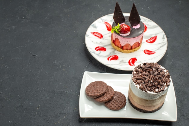 Front view chocolate cake and biscuits on white rectangular plate and cheesecake on white oval plate on dark