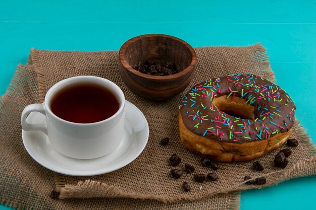 Вид спереди шоколадного пончика с чашкой чая и конфет на бежевой салфетке