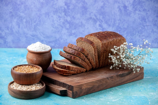 Front view of chopped in half black bread slices on wooden boards flour wheat oatmeal in bowls flower eggs on light ice blue pattern background