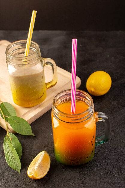 A front view cold cocktails colored inside glass cans with colorful straws green leaves on the wooden cream desk and dark