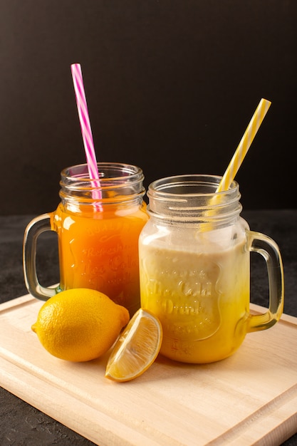 A front view cold cocktails colored inside glass cans with colorful straws on the wooden cream desk and dark