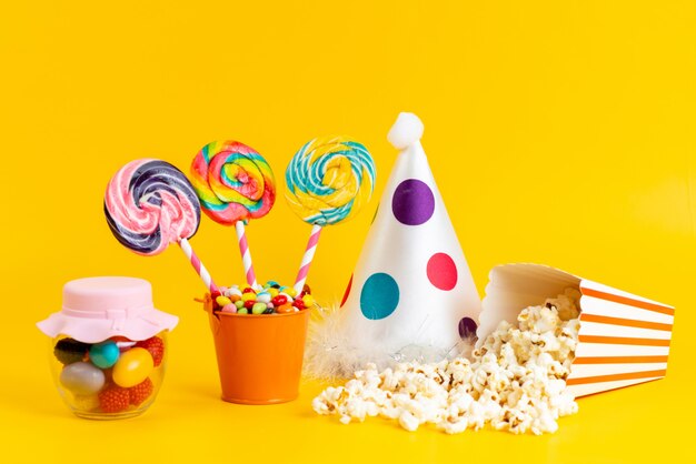 Вид спереди цветные леденцы на палочке с разноцветными конфетами, забавная шапочка и попкорн на желтом