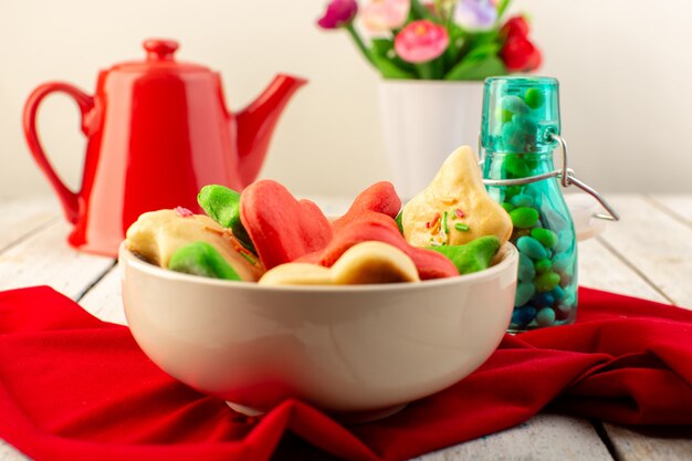 Front view of colorful delicious cookies different formed inside plate with red kettle and candies