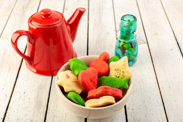 Front view of colorful delicious cookies different formed inside plate with red kettle and candies