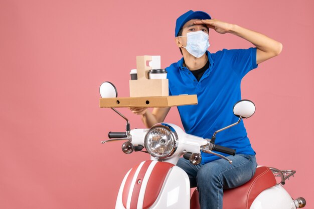 Front view of concentrated male delivery person in mask wearing hat sitting on scooter showing orders on pastel peach background