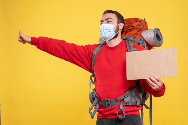 Вид спереди сконцентрированного парня-путешественника в медицинской маске с рюкзаком, показывающий лист без письма на желтом фоне
