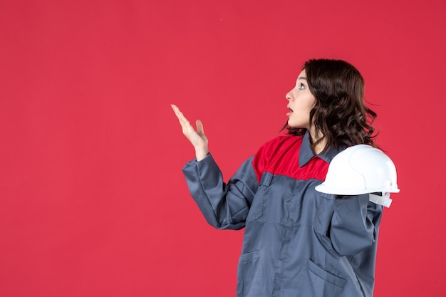 Vista frontale dell'architetto femminile interessato che tiene il cappello duro e rivolto verso l'alto sul lato destro sulla parete rossa isolata