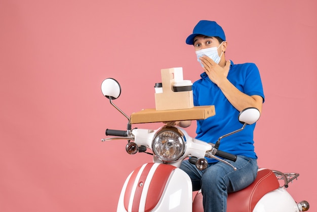 Front view of concerned male delivery person in mask wearing hat sitting on scooter delivering orders on pastel peach background