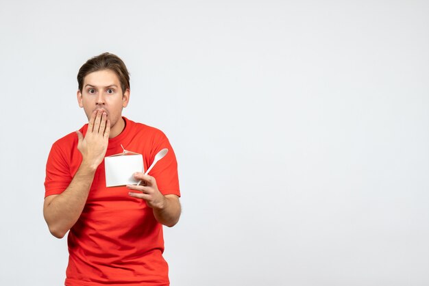 Front view of concerned young guy in red blouse holding paper box and spoon on white background