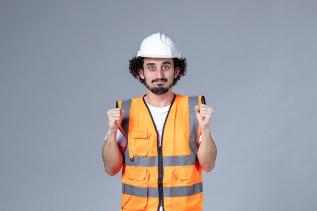 Front view of confident ambitious male builder wearing warning vest with safety helmet on gray wave wall