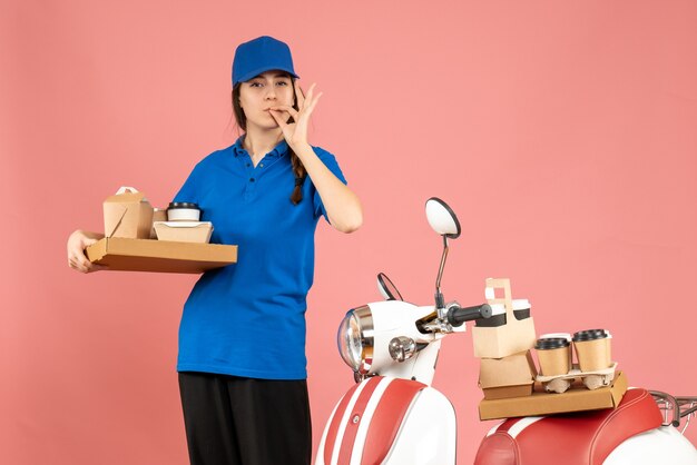 Front view of confident courier girl standing next to motorcycle holding coffee and small cakes on pastel peach color background