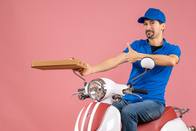 Front view of confident courier man wearing hat sitting on scooter holding order making ok gesture on pastel peach background