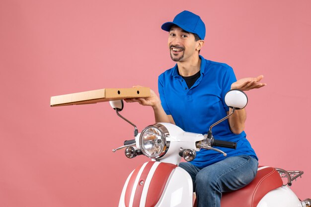 Front view of confident courier man wearing hat sitting on scooter holding order on pastel peach background