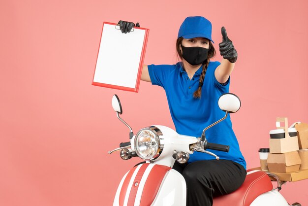 Front view of confident courier woman wearing medical mask and gloves sitting on scooter holding empty paper sheets delivering orders making ok gesture on pastel peach background