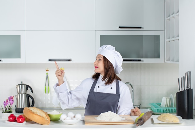 白いキッチンで上向きのまな板パン野菜とテーブルの後ろに立っている制服を着た自信のある女性シェフの正面図