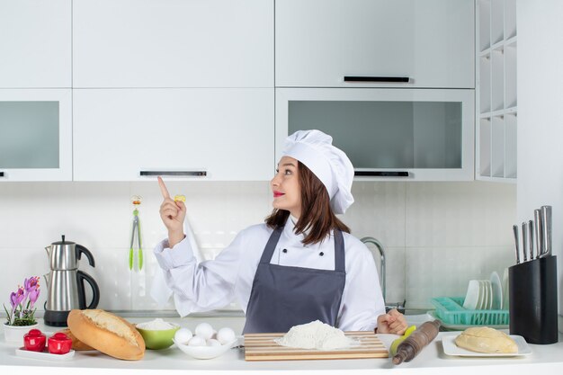 Front view of confident female chef in uniform standing behind the table with cutting board bread vegetables pointing up in the white kitchen