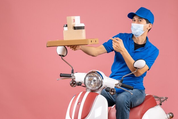 Front view of confident male delivery person in mask wearing hat sitting on scooter delivering orders on pastel peach background