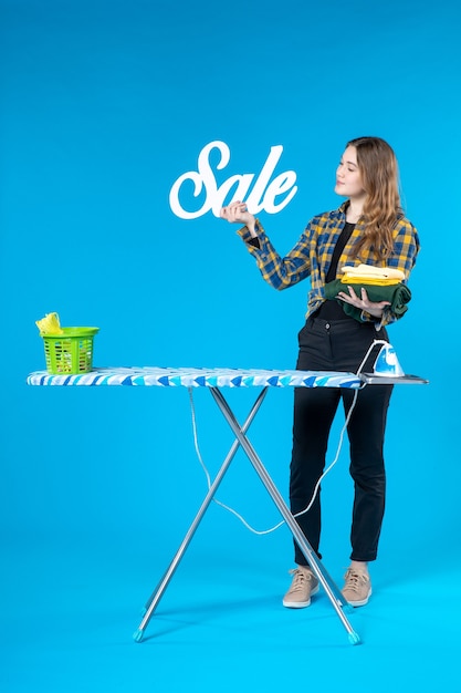 Front view of confident young female holding the folded clothes sale icon and standing behind the ironing board in the laundry room on blue table