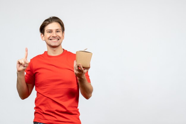 Front view of confident young guy in red blouse holding small box and pointing up on white background