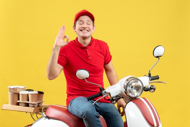 Front view of confident young guy wearing red blouse and hat delivering orders making eyeglasses gesture on yellow background