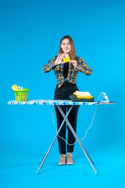 Front view of confident young lady showing bank card and standing behind the ironing board in the laundry room