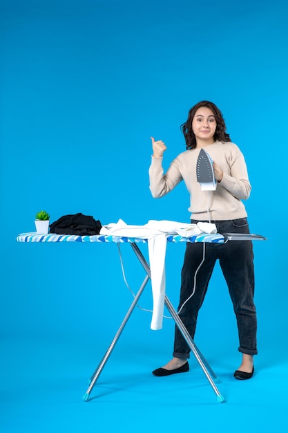 Front view of confident young lady standing behind the board and ironing the clothes pointing up on blue background