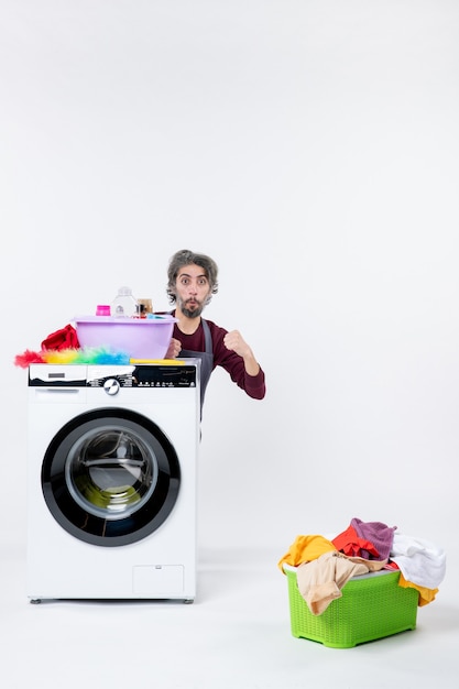 Front view confident young man in apron sitting behind washer laundry basket on white background