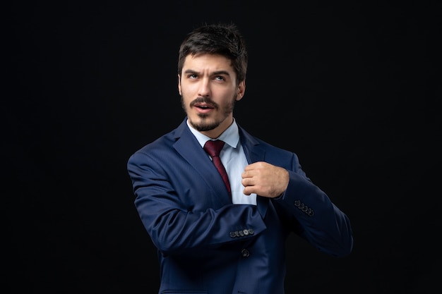 Front view of confused bearded man in suit looking for something in the pocket inside the suit on dark wall