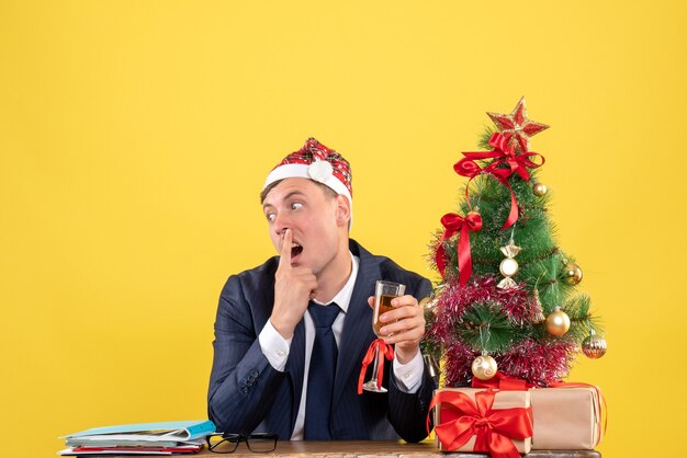 Вид спереди сбитого с толку делового человека, подающего тосты, сидя за столом возле рождественской елки и подарков на желтом