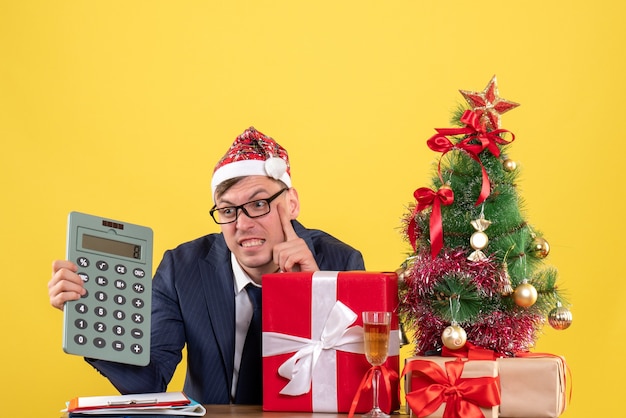 Вид спереди сбитого с толку делового человека с калькулятором, сидящего за столом возле рождественской елки и подарков на желтом