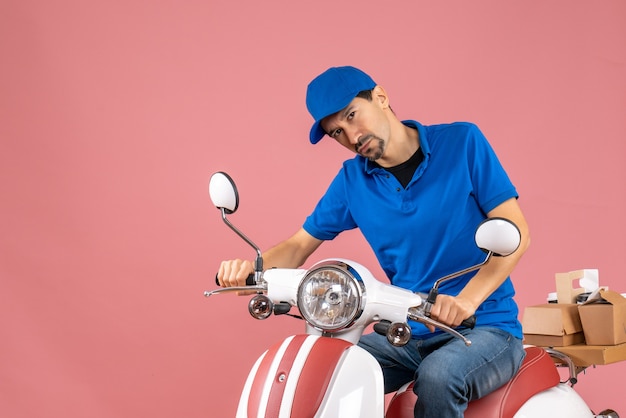 Front view of confused courier guy wearing hat sitting on scooter on pastel peach background