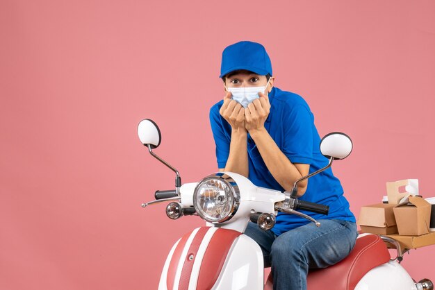 Front view of confused delivery guy in medical mask wearing hat sitting on scooter on pastel peach background