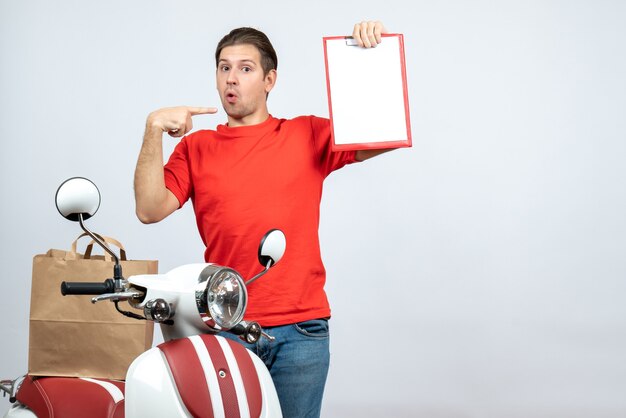 Front view of confused delivery man in red uniform standing near scooter showing document pointing himself on white background