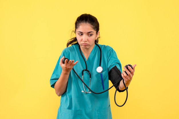 Front view confused female doctor in uniform holding sphygmomanometers standing on yellow background