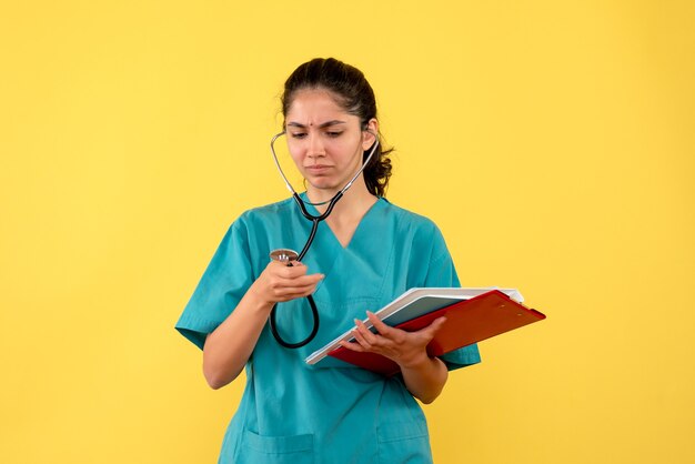 Front view confused female doctor in uniform using stethoscope holding papers on yellow background