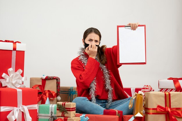 Front view confused girl with black mask holding documents sitting around presents