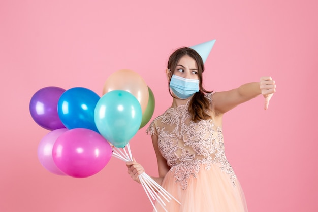 Front view confused girl with party cap and medical mask making thumb down sign holding colorful balloons