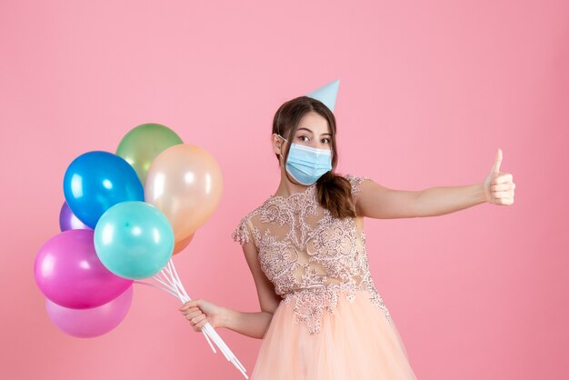 Front view confused girl with party cap and medical mask making thumb up sign holding colorful balloons