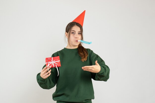 Front view confused girl with party cap pointing at her xmas gift using noisemaker