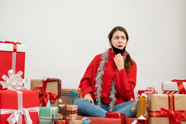 プレゼントの周りに座っている彼女のマスクを脱いで赤いセーターと正面の混乱した女の子